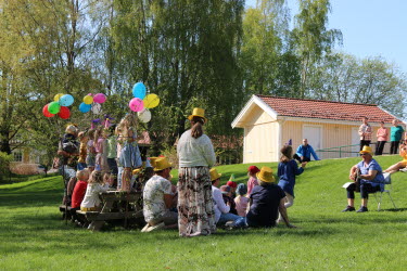 Barn från Böleängens förskola står på rad och sjunger låtar i det gröna gräset. Framför barnen sitter en pedagog och spelar gitarr med en guldig hatt.