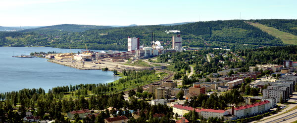 En flygbild över Timrå kommun. Vatten omger en halvö där SCA Östrands fabrik ligger. Skönviksbacken i bakgrunden. Grönskande skog, byggnader och en väg.