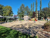En lekplats med sandlåda, gungor och en klätterkub med rutschkanor. Lekparken är omgiven av höga träd, solen skiner och himlen är blå.