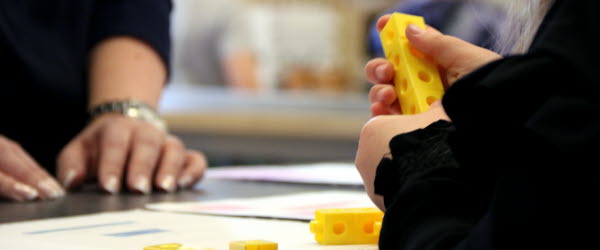 Ett barn håller i gula byggklossar och sitter vid ett bord i ett klassrum. På bordet ligger papper och fler gula byggklossar.