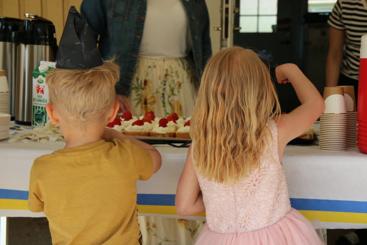 Två barns ryggar som står framför fikabordet och är påbäg att ta varsin sommartårtbit.