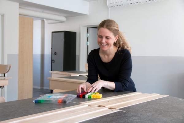 En kvinnlig lärare står i ett klassrum framför ett bord. Hon är glad. På bordet ligger undervisningsmaterial i form av byggbara klossar i olika färger.