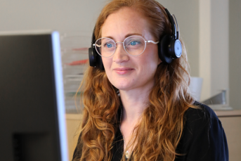 En kvinna med rött lockigt hår och glasögon sitter och arbetar vid en datorskärm. På huvudet har hon svarta hörlurar.