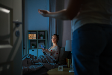Ung kvinna sitter i sängen med mobil och tittar på en person som står bredvid, i ett svagt upplyst rum.