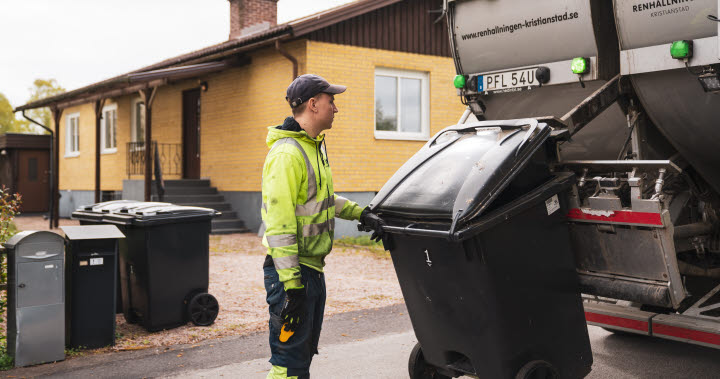 En soptunna lyfts upp av sopbilen för tömning. En person i varselkläder står bredvid.