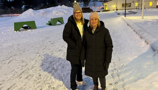 Två kvinnor står på en skolgård och tittar in i kameran och ler. Det är vinter ute.