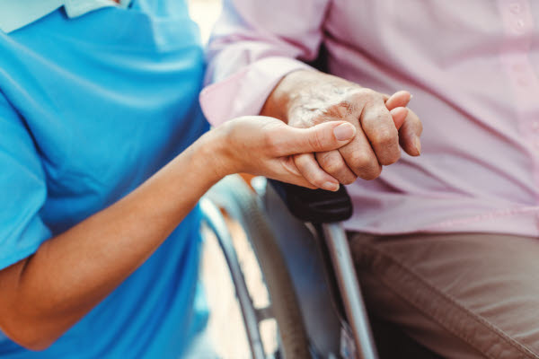 En medarbetare inom vård och omsorg håller handen på en äldre person som sitter i rullstol.