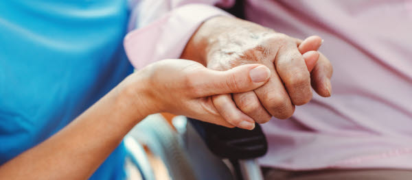 En medarbetare inom vård och omsorg håller handen på en äldre person i rullstol.