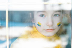 Ett barn tittar ut genom ett fönster. På kinderna har hon målat den ukrainska flaggan.
