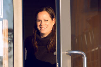 En kvinna öppnar en dörr till en utomhusentré. Hon ler och har solen i ansiktet.