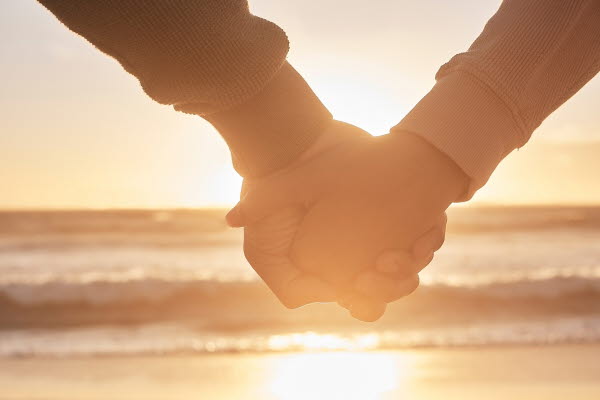 Två personer håller händerna på en strand i solnedgången.