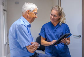 En äldre man i blå skjorta samtalar med en sjuksköterska som håller i en iPad.