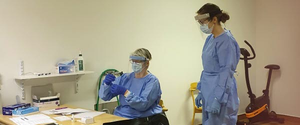 En kvinna sitter vid ett bord och instruerar en annan kvinna i hur antigentestning fungerar. Båda kvinnorna är klädda i blåa arbetskläder och har blåa gummihandskar på händerna. Över ansiktet har de ansiktsmask och visir.