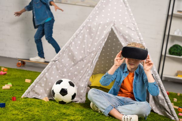 Två barn i ett lekrum. Ett av barnen sitter ner och har på sig VR-glasögon. Andra barnet åker skateboard i bakgrunden.