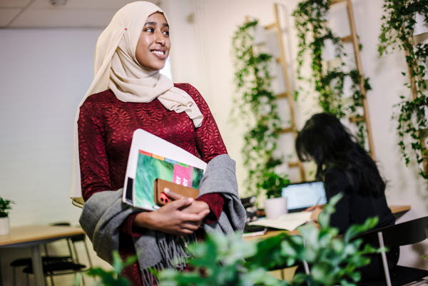 En kvinnlig elev med hijab och böcker i händerna. Gröna växter hänger på väggen.